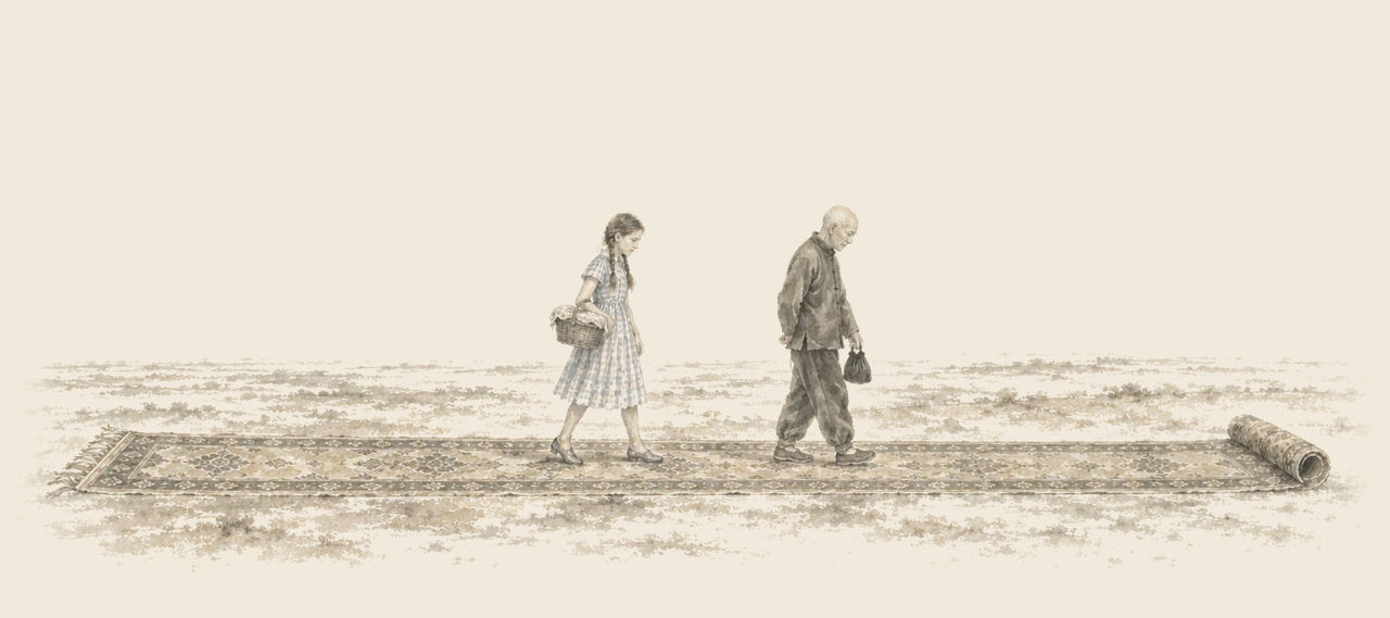 Dorothy, carrying a covered basket, and the Wizard, carrying a small black bag, walk along a carpet unrolling across a desert.